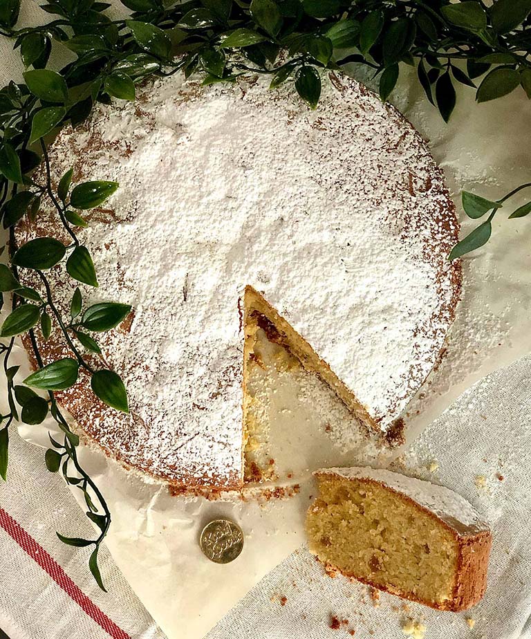 Vasilopita, tradicionalni grčki novogodišnji kolač posut šećerom u prahu, sa izrezanim parčetom koje je prevrnuto pored kolača. Ispod kolača se vidi zlatni novčić sa natpisom 'Kali Xronia', što u prevodu znači 'Srećna Nova Godina'. Kolač je postavljen na belom stoljnjaku sa crvenom trakom i okružen je svežim zelenim listovima, što dodaje svečani ugođaj kompoziciji.