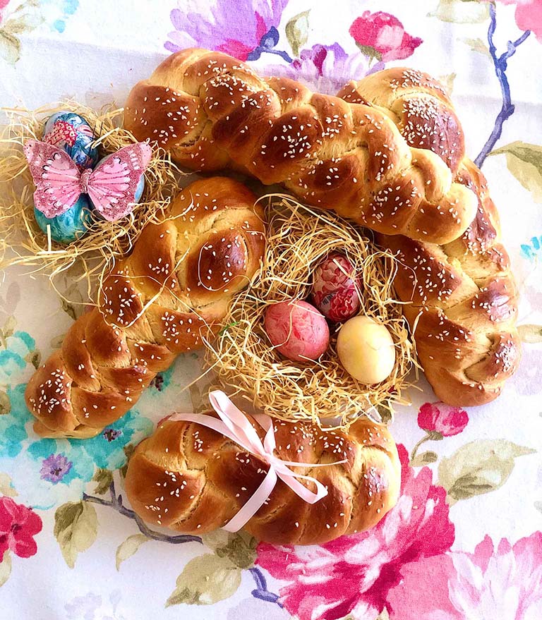 Uskršnji slatki hleb 'tsoureki', u obliku pletenice, posut susamom, sa gnezdima za ptice u centru i šarenim uskršnjim jajima kao i dekorativnim leptirom. Hleb je postavljen na živopisnom cvetnom stolnjaku, stvarajući sliku punu boja i prazničnog duha Uskrsa.