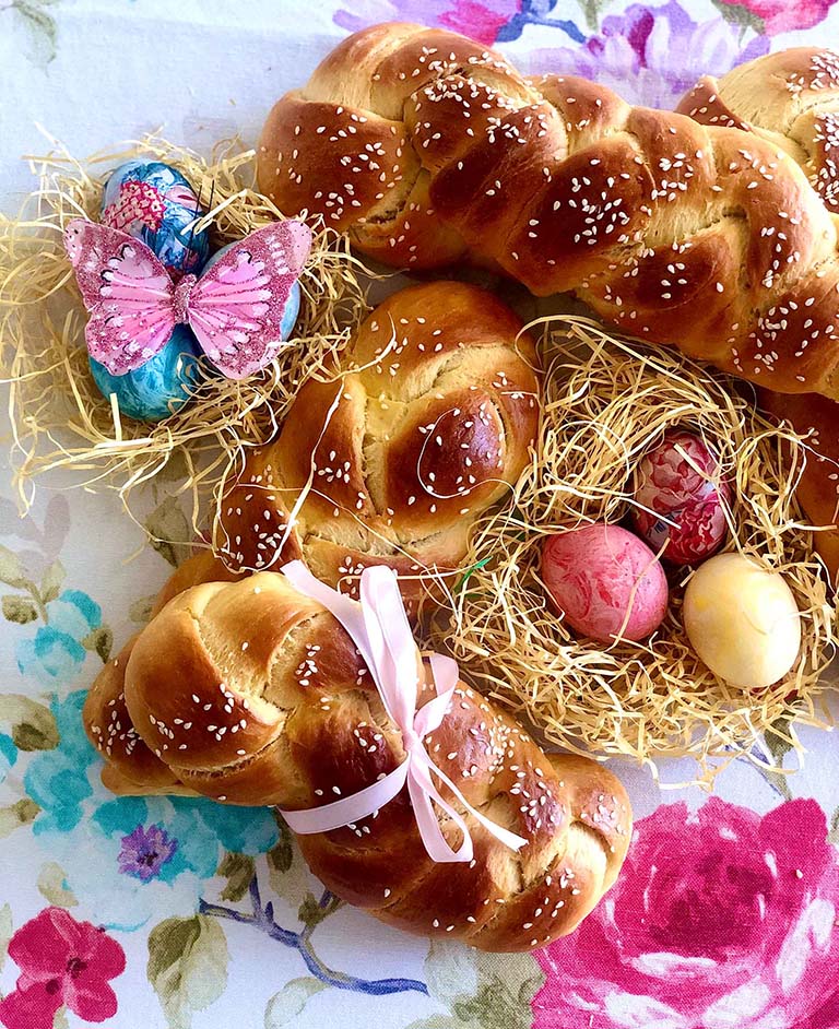 Tradicionalni grčki uskršnji hleb 'tsoureki', svetlo smeđe boje sa sjajnom glazurom i susamom, u obliku pletenica sa roze mašnicom. Pored hleba je gnezdo za ptice od suve trave sa šarenim uskršnjim jajima i ručno oslikanim plavim jajima i dekorativnim leptirom preko. Sve je postavljeno na stolnjaku sa bogatim cvetnim detaljima ističući praznični ambijent i prolećno veselje.