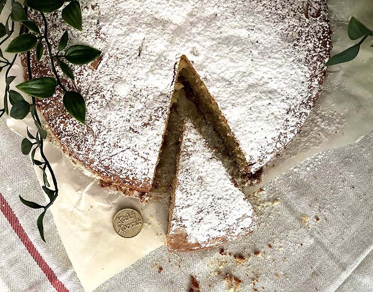Tradicionalna vasilopita torta posuta šećerom u prahu, sa izvađenim jednim parčetom, na belom stolnjaki sa crvenim prugama i dekorativnim zelenim listovima u pozadini. Pored torte se nalazi zlatni novčić sa natpisom 'Kali Xronia', što na grčkom jeziku znači “Srećna Nova Godina”.